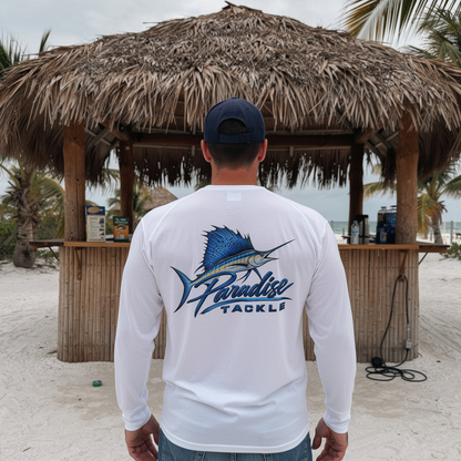 A man standing on a sandy beach facing a thatched-roof beach bar, wearing a white long-sleeve performance fishing shirt with a blue marlin graphic and 'Paradise Tackle' logo on the back, paired with blue jeans and a navy baseball cap.
